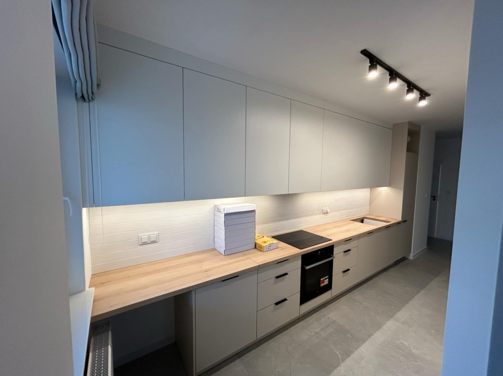 Nowoczesna kuchnia z szarymi szafkami, drewnianym blatem, płytą indukcyjną i piekarnikiem, oświetlona listwą z reflektorami, z pudełkami na blacie.