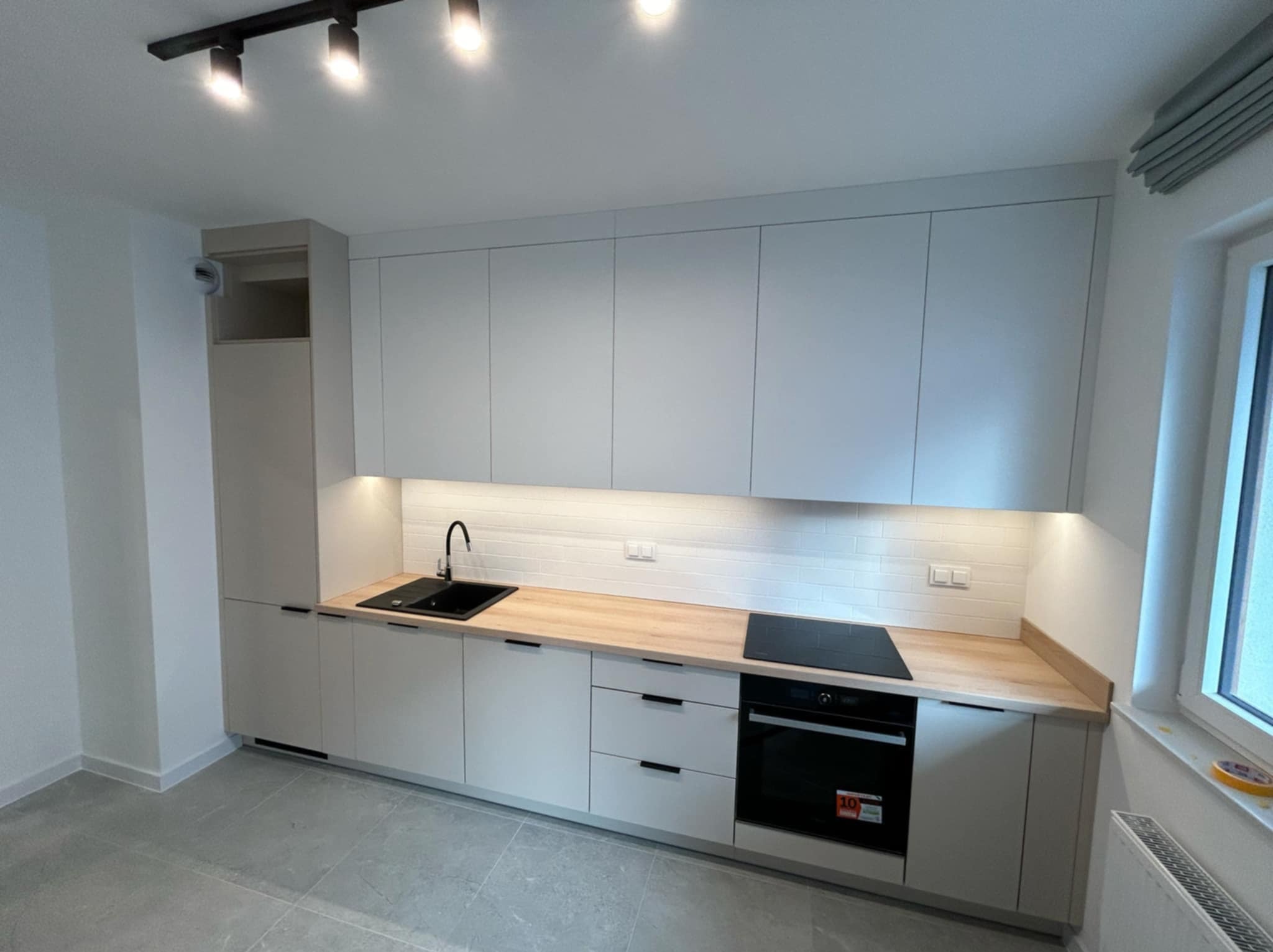 Nowoczesna kuchnia z szarymi szafkami na wymiar, drewnianym blatem, czarnym zlewem i płytą indukcyjną, oświetlona listwą LED pod szafkami wiszącymi.