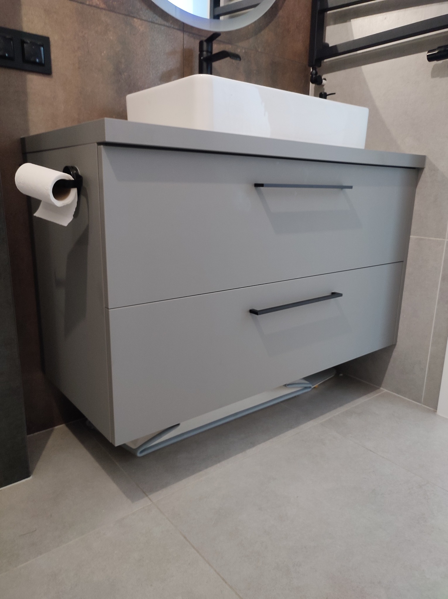 Szara szafka pod umywalkę z dwiema szufladami i czarnymi uchwytami, zamontowana na ścianie w nowoczesnej łazience z beżowymi płytkami, z widocznym fragmentem rolki papieru toaletowego po lewej...