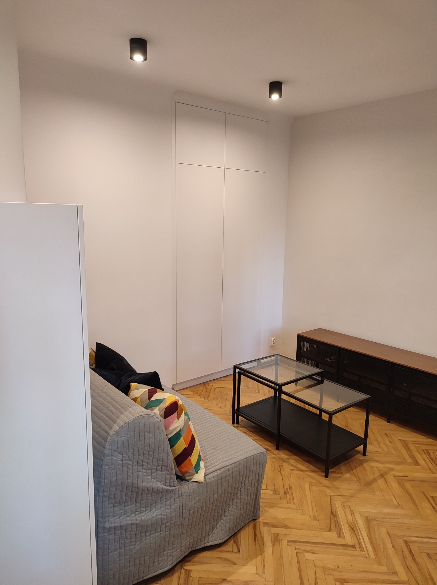 Wnętrze pokoju z wbudowaną białą szafą sięgającą sufitu, szarą sofą z kolorową poduszką, stolikami kawowymi ze szklanym blatem i komodą z ciemnego drewna na parkiecie w jodełkę.