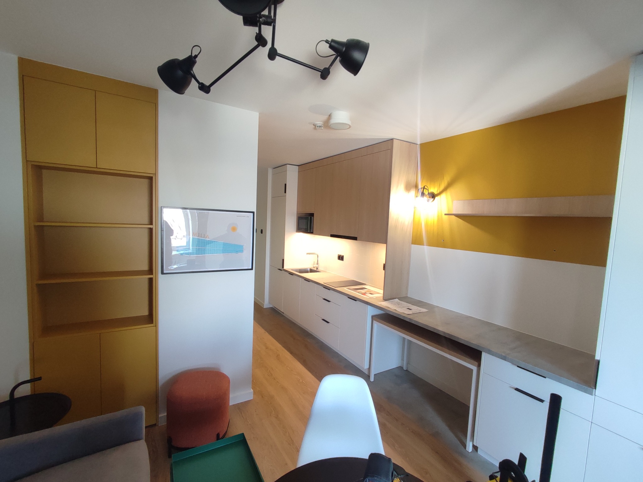 Wnętrze mieszkania z zabudową kuchenną w jasnych odcieniach drewna i bieli, połączoną z biurkiem, oraz żółtą szafą z otwartymi półkami po lewej stronie, oświetlone nowoczesną lampą sufitową.
