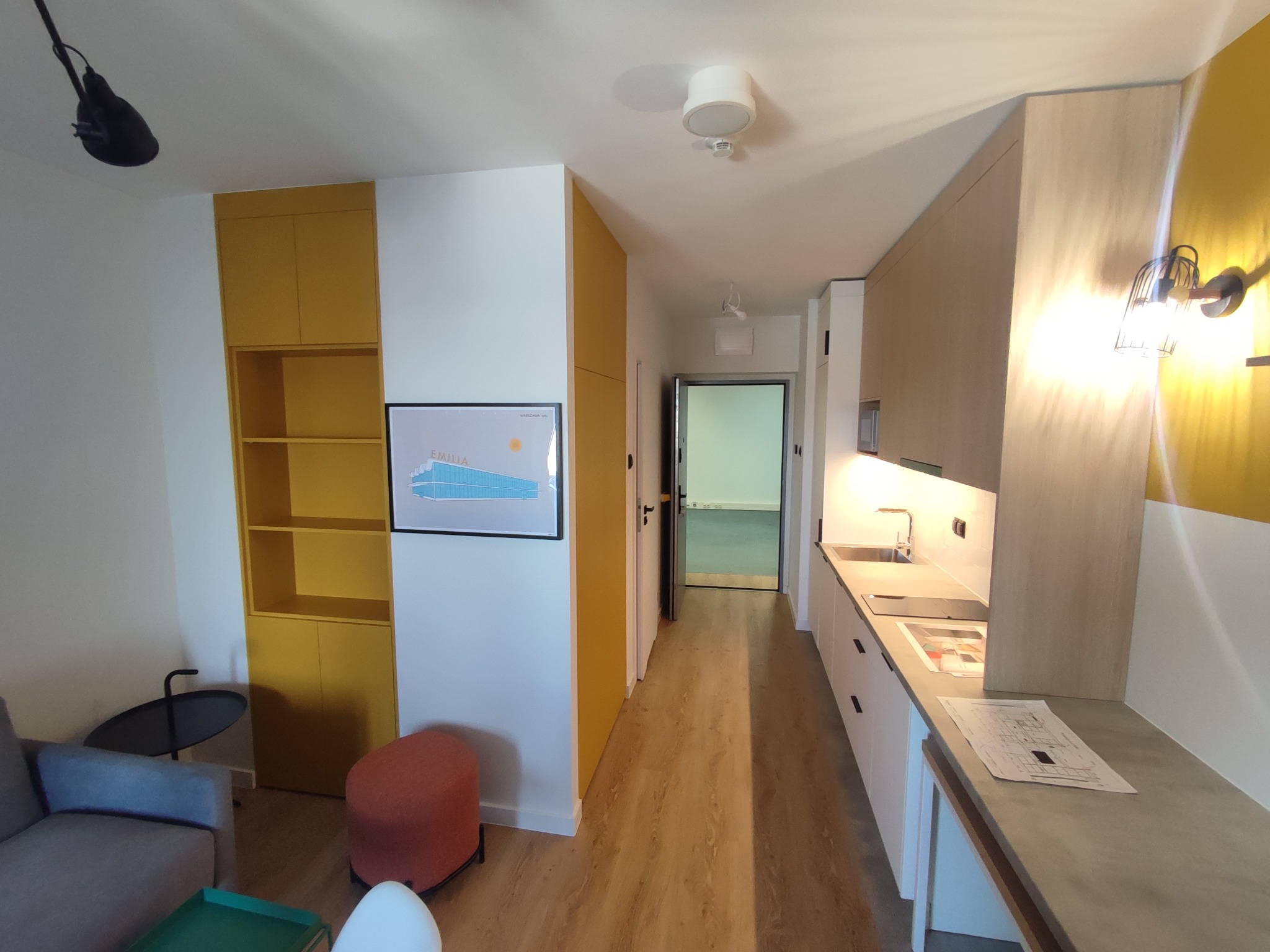 Wnętrze mieszkania z zabudową kuchenną i szafą wnękową w odcieniach żółci i beżu. Na blacie kuchennym leżą plany architektoniczne.