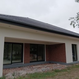 Vitalii Chabaniuk - Nowoczesny dom parterowy z białymi ścianami i ciemną dachówką. Widoczne duże okna i fragment elewacji wykończony czerwoną cegłą. Teren wokół domu w trakcie prac wykończeniowych.
