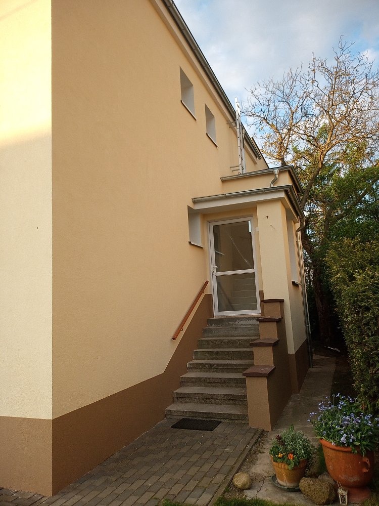 Jasna elewacja budynku z beżowym odcieniem, ciemniejszy pas u dołu, schody prowadzące do przeszklonych drzwi wejściowych, widoczne rośliny w donicach.