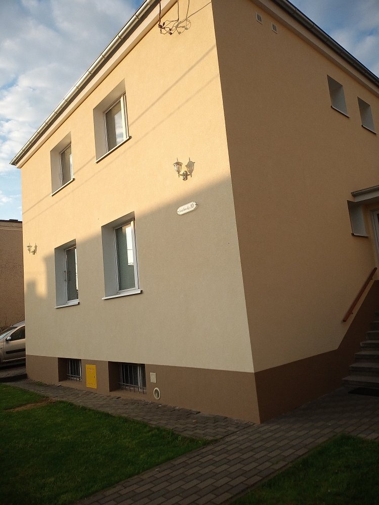 Dwukondygnacyjny budynek z elewacją w odcieniach beżu i brązu, widoczne okna z białymi ramami, lampa zewnętrzna i tabliczka z numerem domu, trawnik i chodnik z kostki brukowej.