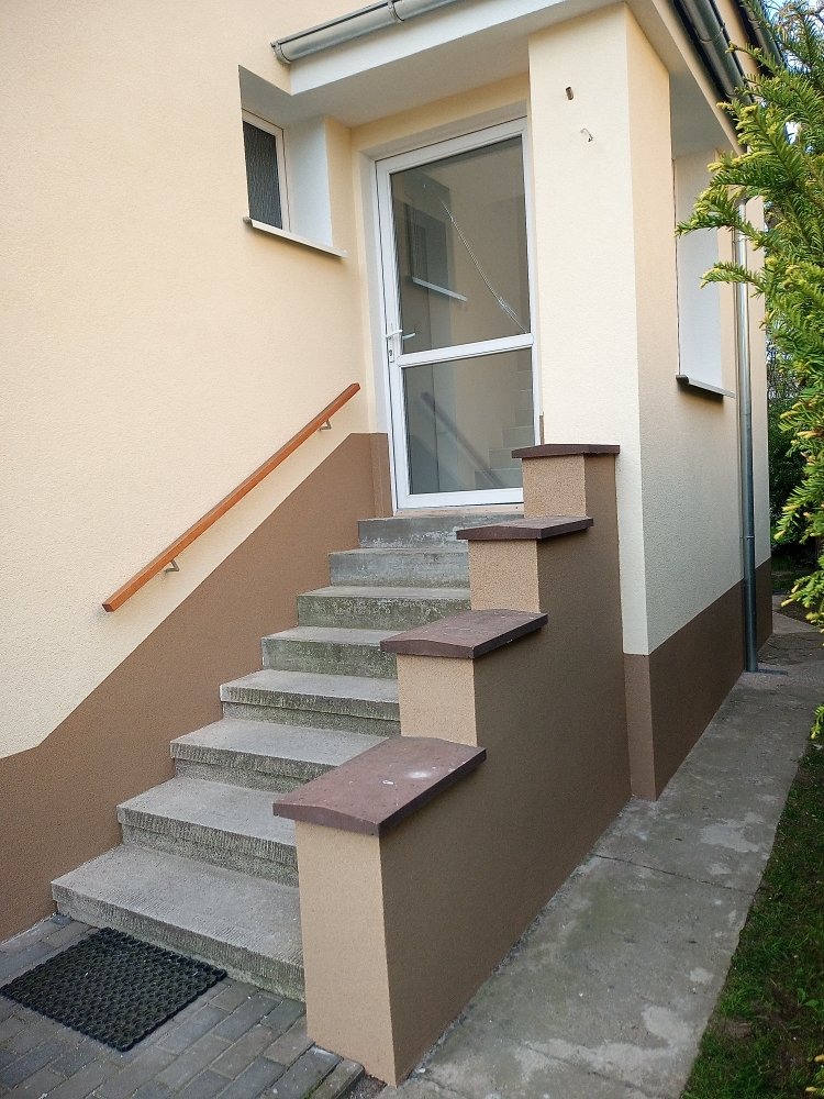 Dwukolorowa elewacja domu z beżowo-brązowym malowaniem, schody wejściowe z szarego betonu, białe drzwi wejściowe z szybą i drewniana poręcz