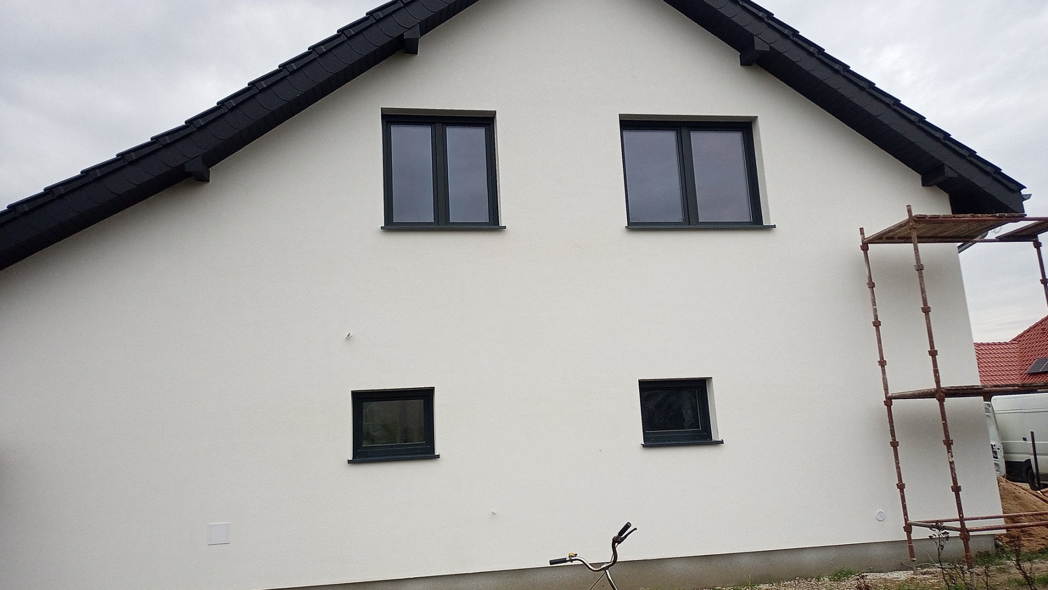 Świeżo otynkowana i pomalowana na biało ściana dwukondygnacyjnego domu z czarną dachówką i ramami okiennymi, widoczne rusztowanie z boku budynku.