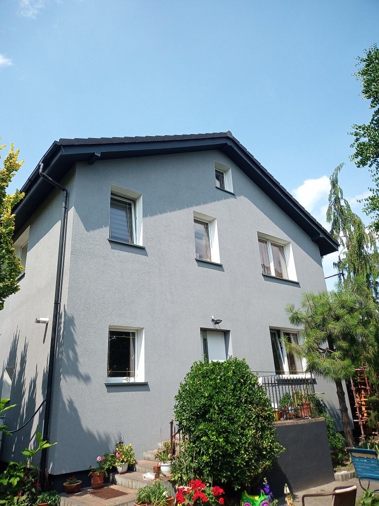 Dwupiętrowy dom z szarą elewacją, czarnym dachem i białymi ramami okiennymi, widoczny z dołu w słoneczny dzień. Na pierwszym planie roślinność i schody.