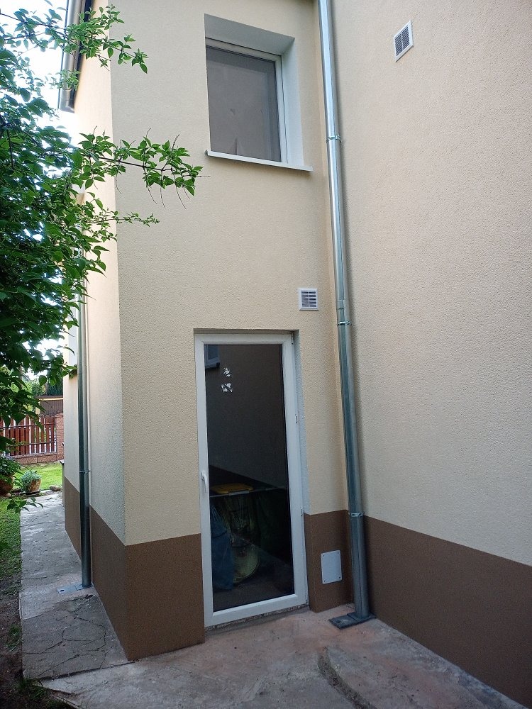 Fragment elewacji budynku z widocznym podziałem kolorystycznym: beżowa góra i brązowy dół, rynna spustowa, okno i białe drzwi wejściowe.