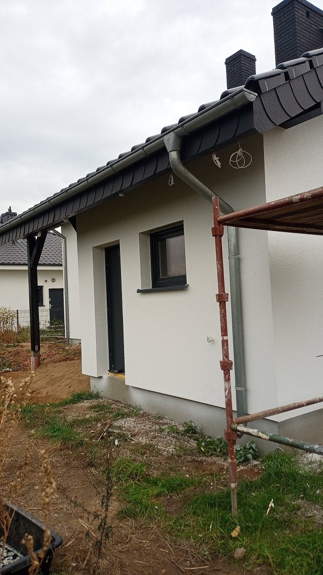 Świeżo otynkowana elewacja domu jednorodzinnego w kolorze białym, z ciemną stolarką okienną i drzwiami wejściowymi, widoczne rusztowanie przy ścianie, dach pokryty ciemną dachówką