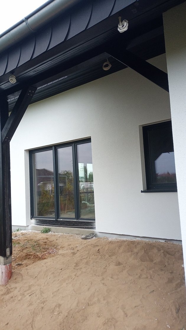 Biała elewacja domu jednorodzinnego z czarnymi ramami okiennymi, widoczna z poziomu gruntu, z fragmentem drewnianej konstrukcji dachu i piaskiem na pierwszym planie.