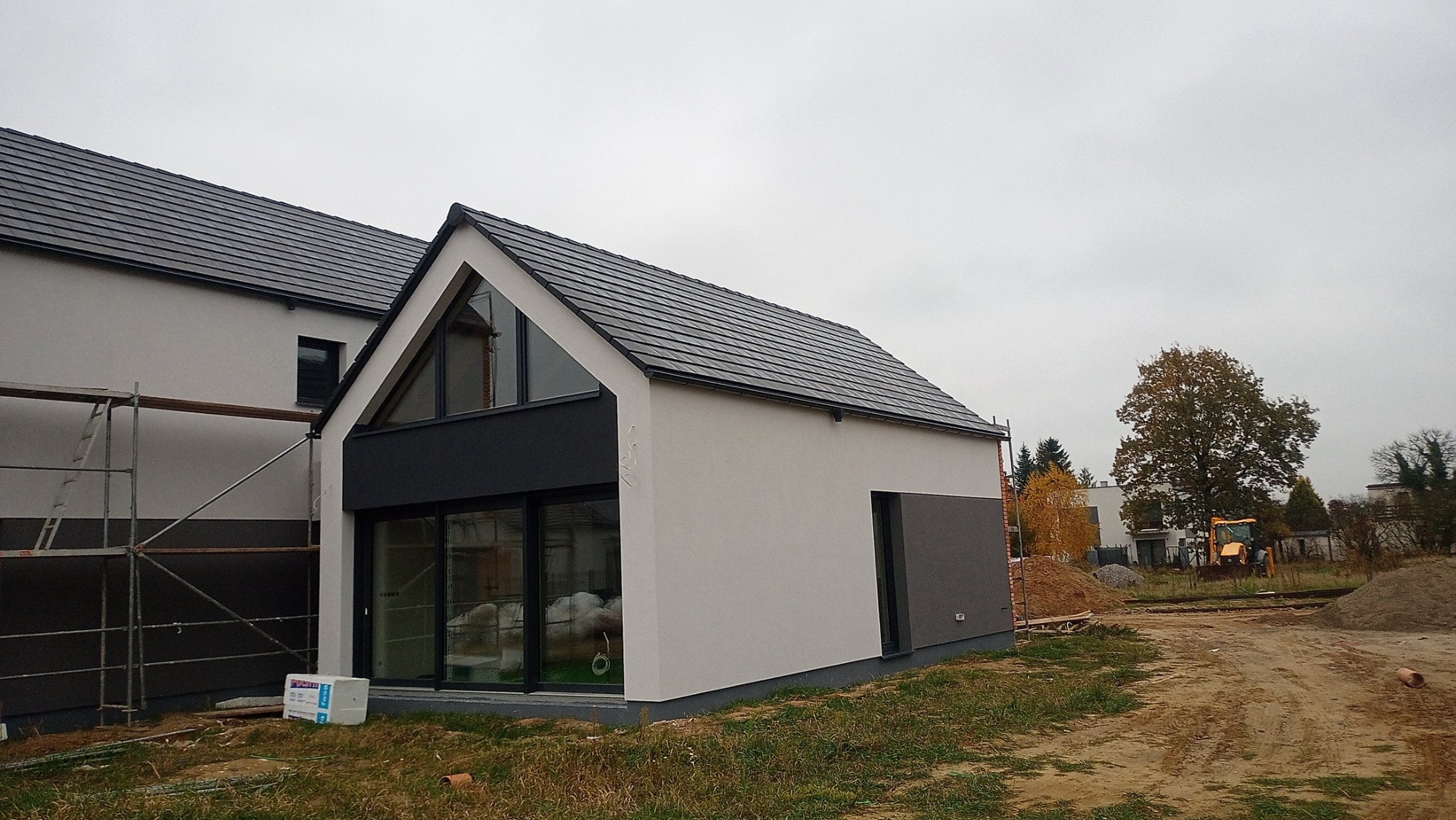 Nowoczesny dom jednorodzinny z szarą elewacją i dużymi oknami, częściowo w trakcie wykańczania, widoczne rusztowania i materiały budowlane na trawniku, w tle koparka.