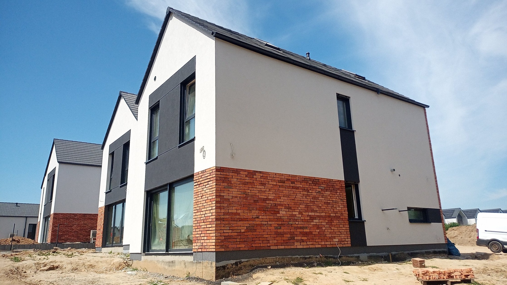 Nowoczesny dom jednorodzinny z elewacją w odcieniach bieli, szarości i cegły, widoczny z perspektywy narożnej, z fragmentem terenu budowy i niebieskim niebem w tle.