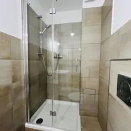 Nowoczesna łazienka z kabiną prysznicową typu walk-in, wykończona płytkami w odcieniach beżu i szarości, widoczny fragment toalety.