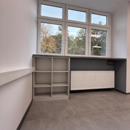 ADAMCO Wykończenia - Szara zabudowa meblowa pod oknem z grzejnikiem i półkami. Widok na zewnątrz przez okno. Szare płytki na podłodze. Minimalistyczny design.