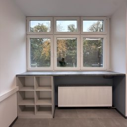 ADAMCO Wykończenia - Białe okno PCV z parapetem i szafką wnękową, widok na jesienne drzewa. Szary grzejnik pod oknem. Minimalistyczne wnętrze z szarą podłogą.