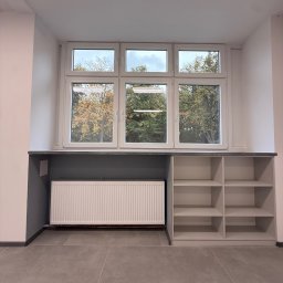 ADAMCO Wykończenia - Białe okno PCV z widokiem na drzewa, pod nim szara zabudowa z grzejnikiem i półkami. Minimalistyczny design, szare płytki podłogowe, neutralna kolorystyka.