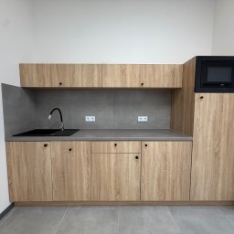 ADAMCO Wykończenia - Jasna, drewnopodobna zabudowa kuchenna z czarnym zlewem i mikrofalówką. Minimalistyczny design, szare płytki na ścianie i podłodze. Nowoczesna kuchnia.