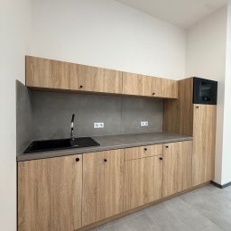 ADAMCO Wykończenia - Jasna kuchnia z drewnianymi szafkami i szarym blatem. Czarny zlew i bateria. Minimalistyczny design, nowoczesne wykończenie, widoczne gniazdka elektryczne.