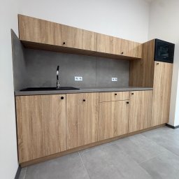 ADAMCO Wykończenia - Jasna kuchnia na wymiar z drewnianymi frontami i szarym blatem. Minimalistyczny design, czarne okrągłe uchwyty, szara ściana nad blatem. Nowoczesne wykończenie.