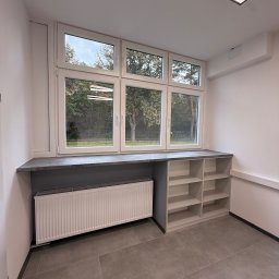ADAMCO Wykończenia - Wnętrze pokoju po remoncie: okno PCV z widokiem na zieleń, grzejnik pod parapetem, szara zabudowa z półkami i szare płytki podłogowe. Minimalistyczny design, neutralna kolorystyka.