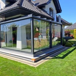 ADAMCO Wykończenia - Nowoczesna, przeszklona pergola metalowa przylegająca do domu z zadbanym trawnikiem. Widoczne detale wykończenia i osoba wewnątrz konstrukcji.