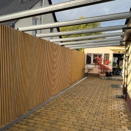 ADAMCO Wykończenia - Nowoczesne ogrodzenie z drewnianych paneli pod metalową pergolą, widok z brukowanego podwórka. Minimalistyczny design, pionowe linie, ciepłe odcienie drewna.
