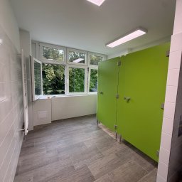 ADAMCO Wykończenia - Nowoczesna toaleta publiczna z zielonymi kabinami, szarą podłogą imitującą drewno i dużym oknem z widokiem na zieleń. Minimalistyczny design, jasne oświetlenie.