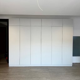 ADAMCO Wykończenia - Biała, minimalistyczna szafa wnękowa z siedmioma drzwiami, na tle szarej podłogi imitującej drewno. Widoczny fragment instalacji elektrycznej na suficie.