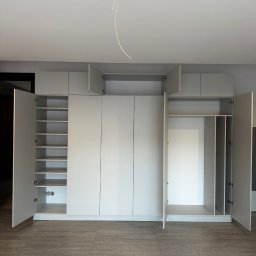ADAMCO Wykończenia - Jasnoszara szafa na wymiar z otwartymi drzwiami, ukazująca półki i przestrzeń do wieszania ubrań. Widoczne szare płytki podłogowe i wystający kabel z sufitu.