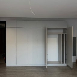 ADAMCO Wykończenia - Jasnoszara szafa na wymiar z otwartymi drzwiami, ukazująca półki i przestrzeń do wieszania. Minimalistyczny design, widoczne płytki podłogowe imitujące drewno.