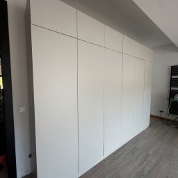 ADAMCO Wykończenia - Biała szafa wnękowa z gładkimi frontami, sięgająca sufitu, na tle szarej podłogi z płytek imitujących kamień. Minimalistyczny design, widoczne detale wykończenia.