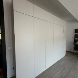 Biała szafa wnękowa z gładkimi frontami, sięgająca sufitu, na tle szarej podłogi z płytek imitujących kamień. Minimalistyczny design, widoczne detale wykończenia.