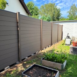 ADAMCO Wykończenia - Nowoczesne ogrodzenie panelowe w kolorze brązowym, z widocznymi słupkami i betonową podmurówką. W tle budynek i błękitne niebo. Ujęcie z poziomu gruntu.