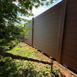 ADAMCO Wykończenia - Nowoczesne ogrodzenie panelowe z kompozytu drewna, widok z dołu, z elementami roślinności i niebem w tle. Brązowe panele z wyraźną fakturą drewna.