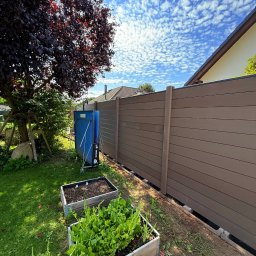 ADAMCO Wykończenia - Nowoczesne ogrodzenie panelowe z kompozytu w kolorze brązowym, widok z ogrodu z grządkami warzywnymi i błękitnym niebem w tle. Estetyczne wykończenie posesji.