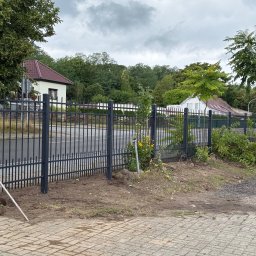 ADAMCO Wykończenia - Ciemne, metalowe ogrodzenie panelowe, częściowo zamontowane, z widocznym wykopem i podporą, na tle domów i zieleni w pochmurny dzień.