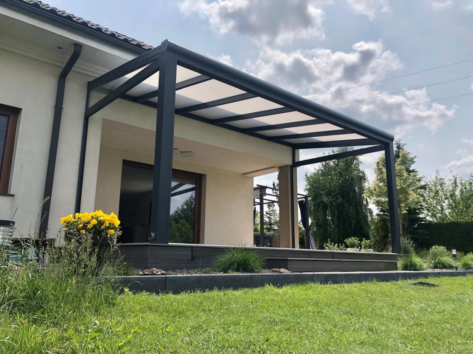 Nowoczesna pergola metalowa z białym zadaszeniem, przylegająca do jasnego budynku. Widoczne kwiaty w donicy i zadbany trawnik. Ujęcie z poziomu trawy.