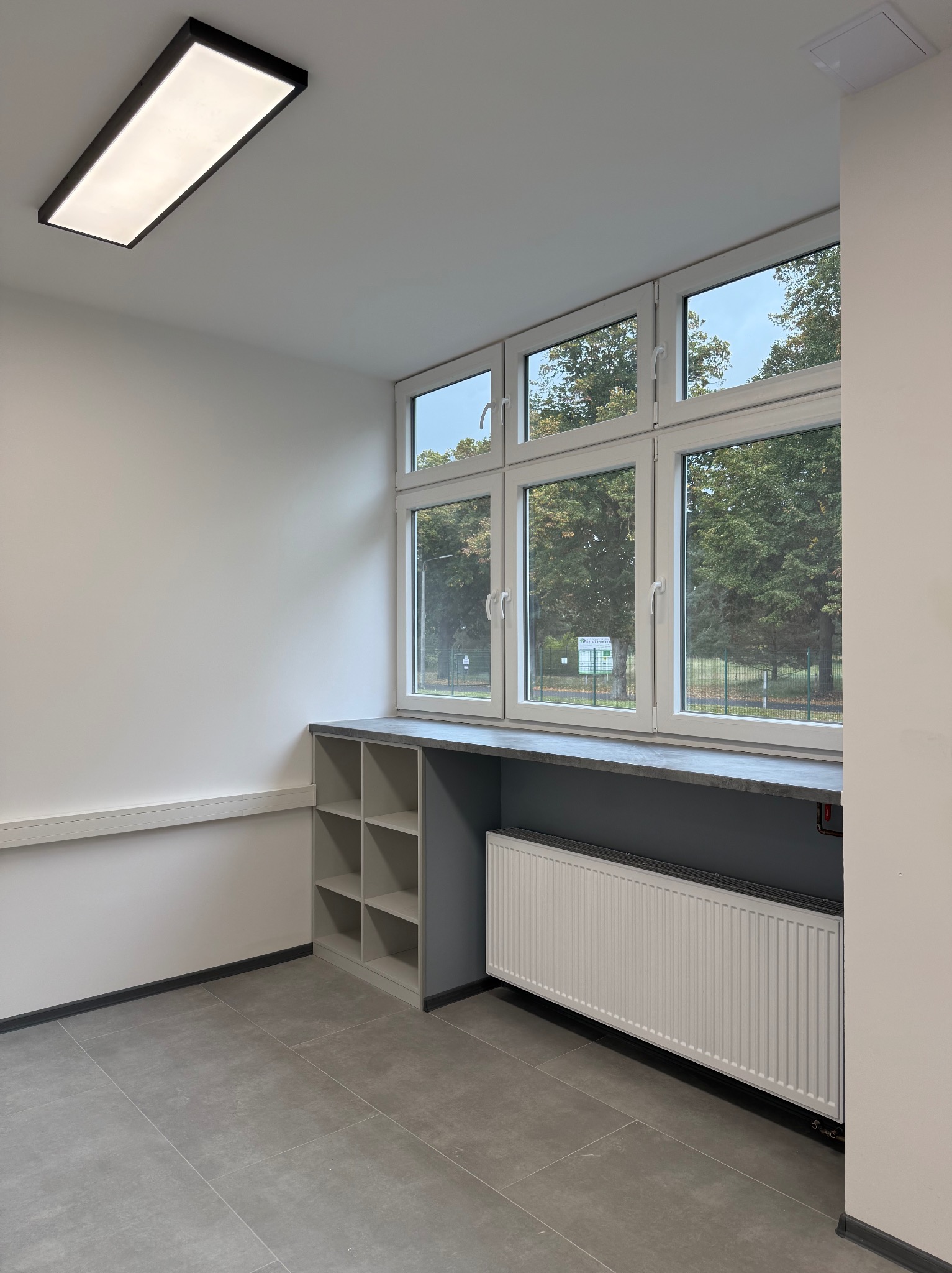 Jasne wnętrze z oknem, grzejnikiem i półkami w szarościach. Widok na zieleń za oknem. Minimalistyczny design, płytki podłogowe i prostokątna lampa sufitowa.