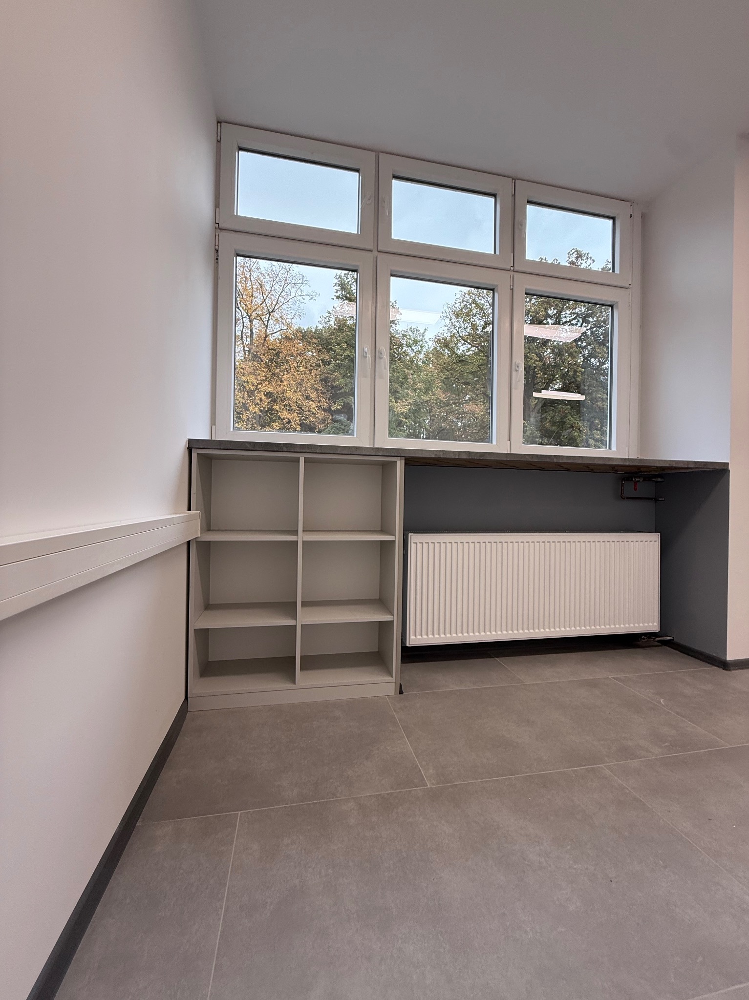 Szara zabudowa meblowa pod oknem z grzejnikiem i półkami. Widok na zewnątrz przez okno. Szare płytki na podłodze. Minimalistyczny design.