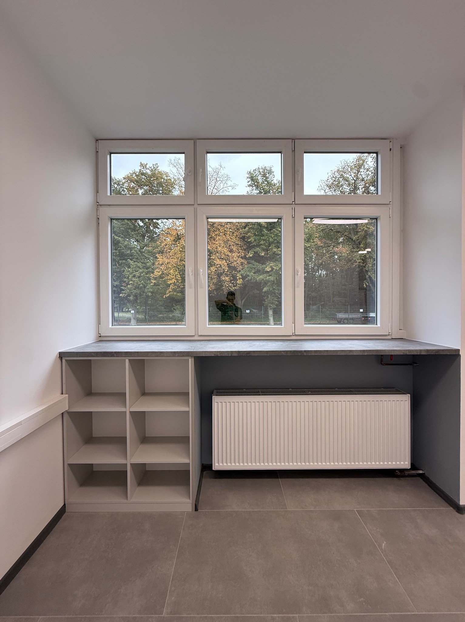 Białe okno PCV z parapetem i szafką wnękową, widok na jesienne drzewa. Szary grzejnik pod oknem. Minimalistyczne wnętrze z szarą podłogą.