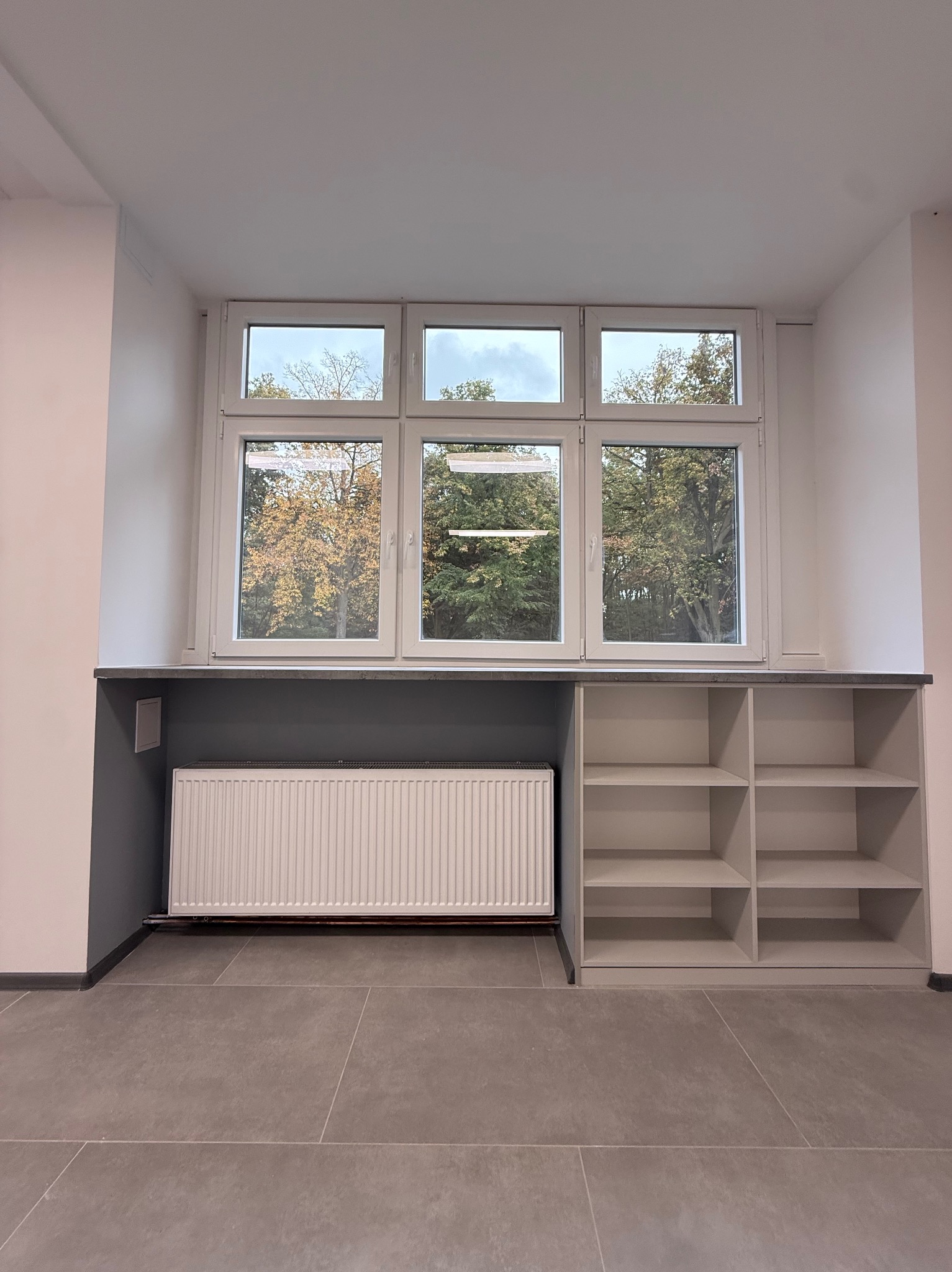 Białe okno PCV z widokiem na drzewa, pod nim szara zabudowa z grzejnikiem i półkami. Minimalistyczny design, szare płytki podłogowe, neutralna kolorystyka.