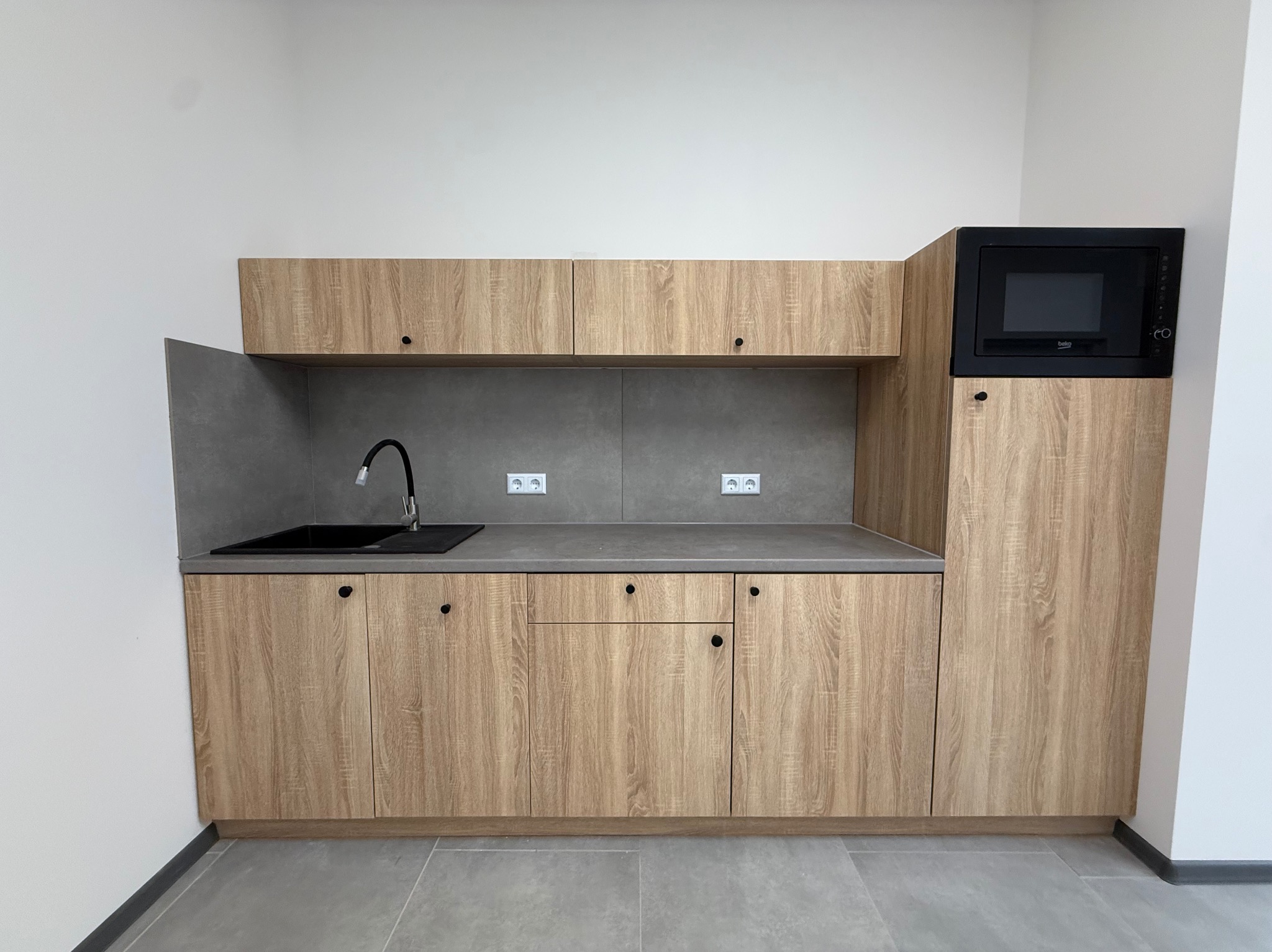 Jasna, drewnopodobna zabudowa kuchenna z czarnym zlewem i mikrofalówką. Minimalistyczny design, szare płytki na ścianie i podłodze. Nowoczesna kuchnia.