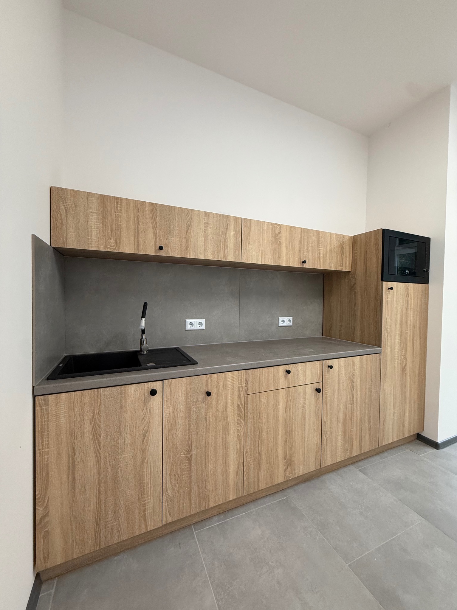 Jasna kuchnia z drewnianymi szafkami i szarym blatem. Czarny zlew i bateria. Minimalistyczny design, nowoczesne wykończenie, widoczne gniazdka elektryczne.