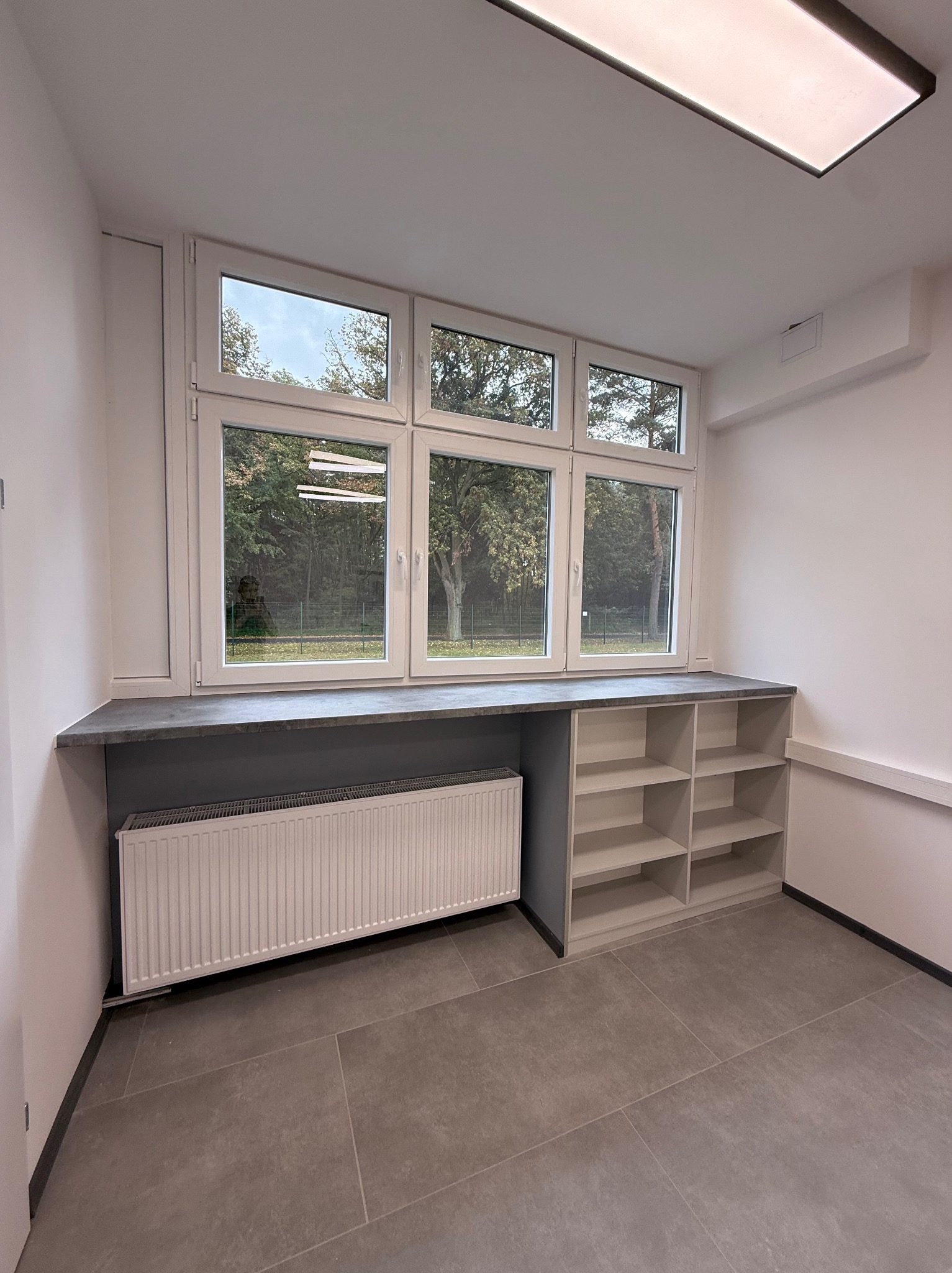 Wnętrze pokoju po remoncie: okno PCV z widokiem na zieleń, grzejnik pod parapetem, szara zabudowa z półkami i szare płytki podłogowe. Minimalistyczny design, neutralna kolorystyka.