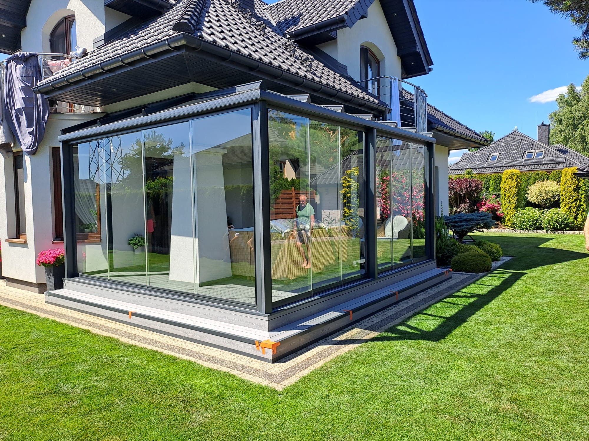 Nowoczesna, przeszklona pergola metalowa przylegająca do domu z zadbanym trawnikiem. Widoczne detale wykończenia i osoba wewnątrz konstrukcji.