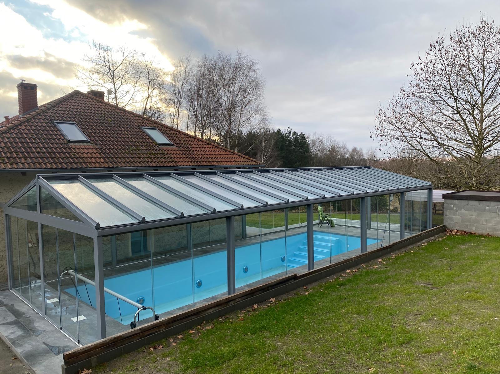 Zadaszony basen z szarą konstrukcją i przezroczystymi ścianami, widoczny z zewnątrz, na tle domu z czerwoną dachówką i zielonego trawnika.