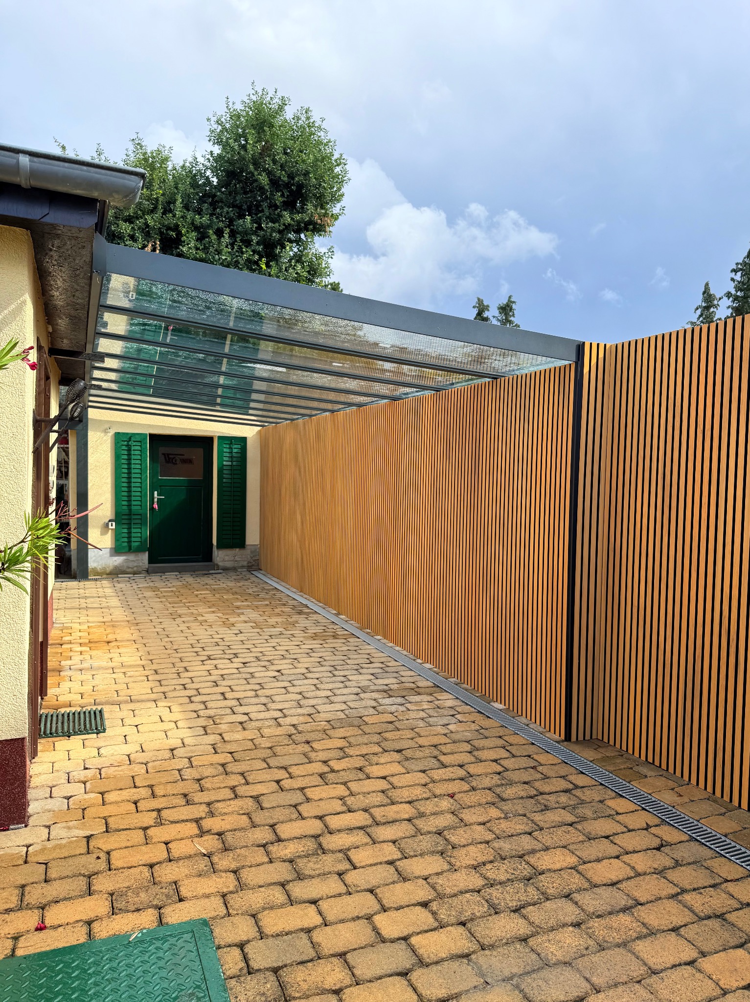 Nowoczesna pergola z drewnianą ścianą lamelową i szklanym zadaszeniem, widok z brukowanej ścieżki. Zielone okiennice i drzwi w tle. Minimalistyczny design.