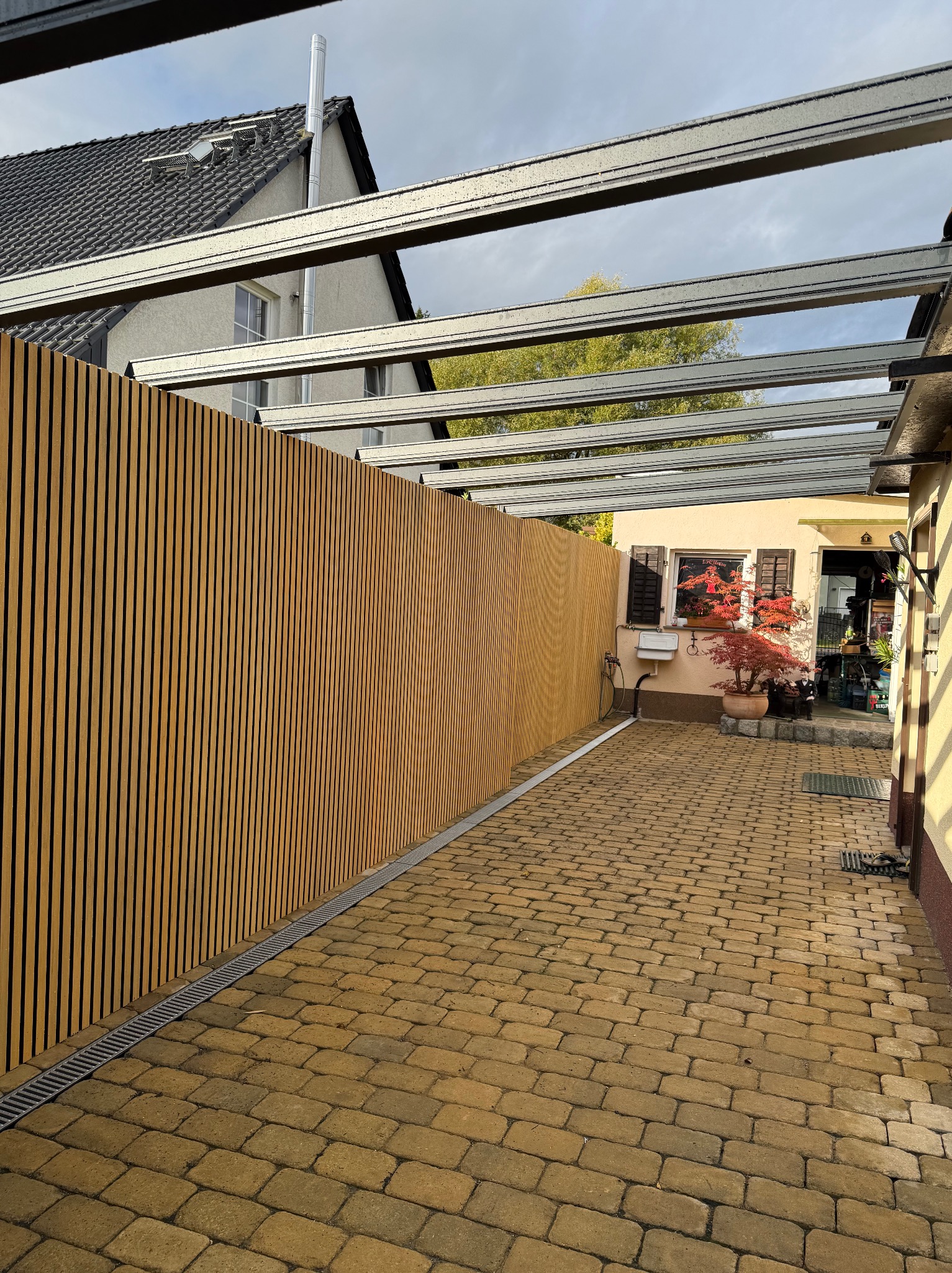 Nowoczesne ogrodzenie z drewnianych paneli pod metalową pergolą, widok z brukowanego podwórka. Minimalistyczny design, pionowe linie, ciepłe odcienie drewna.