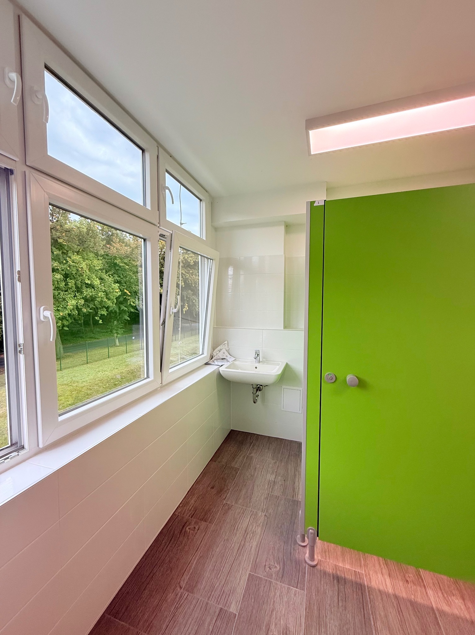 Jasne wnętrze łazienki z oknem PCV, umywalką i zielonymi drzwiami. Widoczne drewnopodobne panele podłogowe i białe płytki ścienne. Częściowo otwarte okno z widokiem na zieleń.